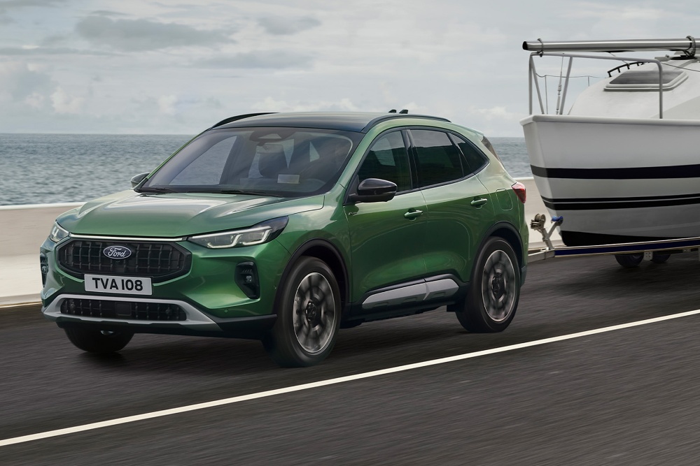 A green Ford Kuga towing a small boat along a coastal road.