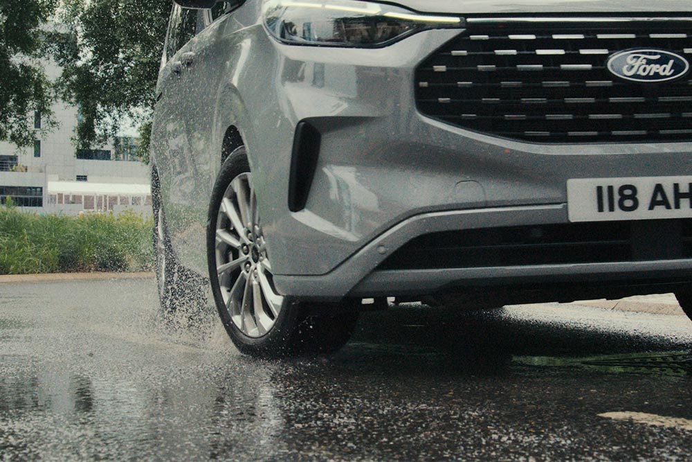 The front wheel of a silver Ford Tourneo Custom splashes through a puddle.