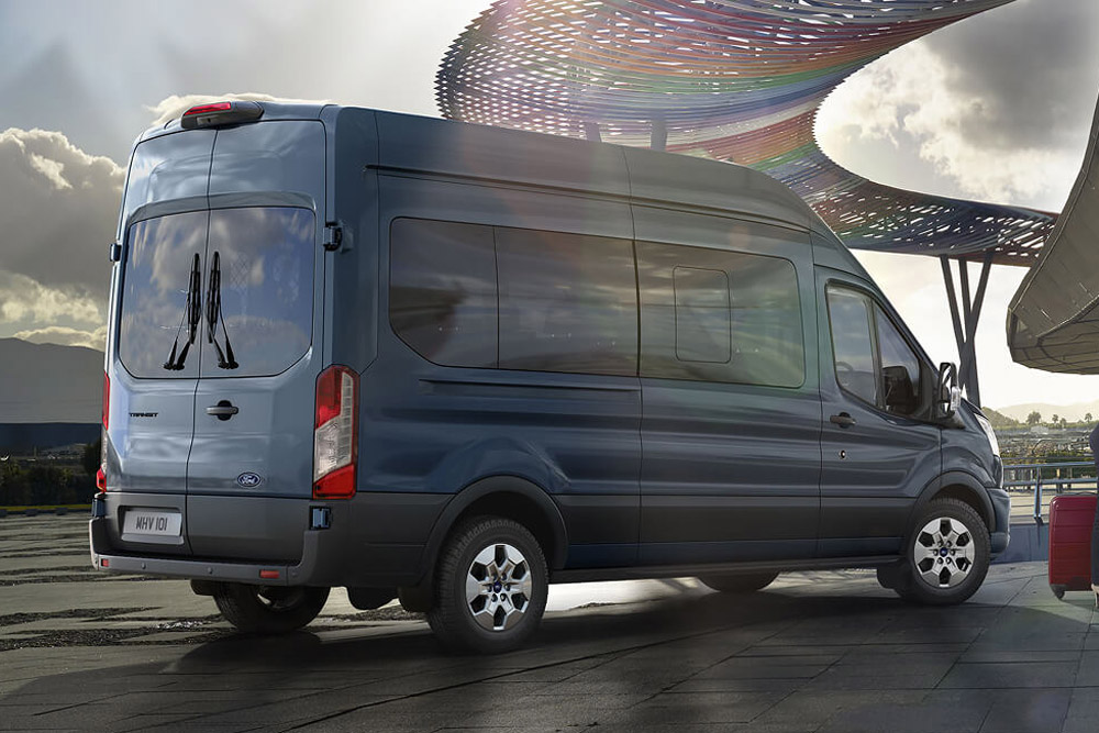 Rear 3/4 view of a blue Ford Transit Minibus parked outdoors, with a modern, colorful structure in the background.