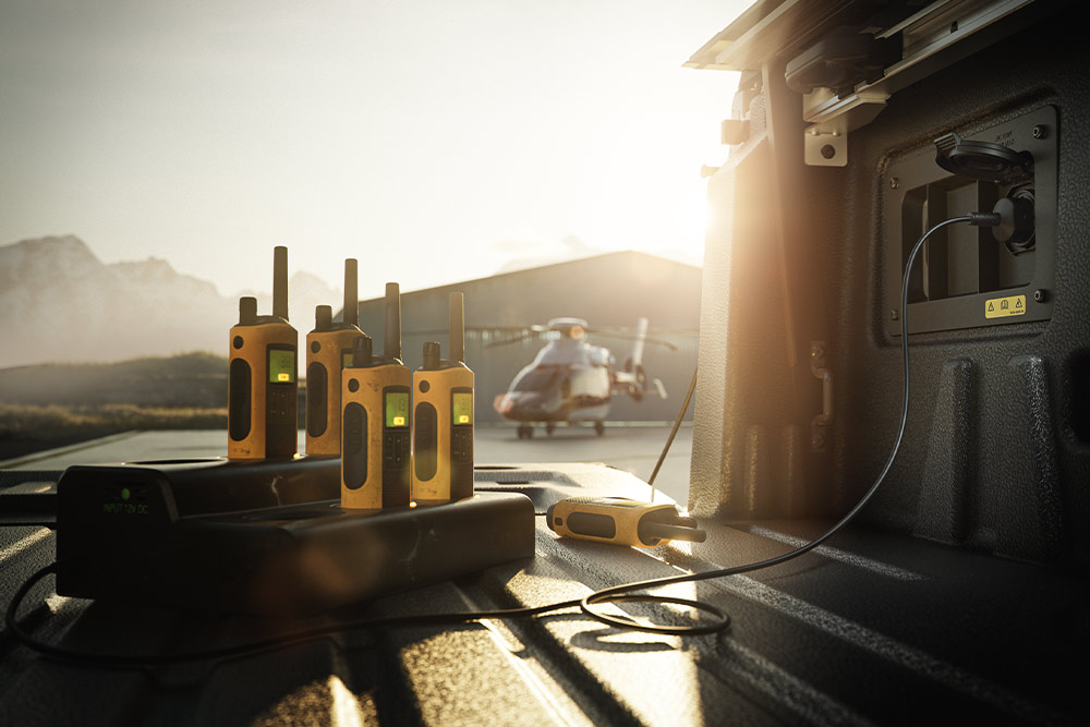 Several walkie-talkies charging in the bed of a Ranger Raptor at sunset, with a helicopter in the distance.