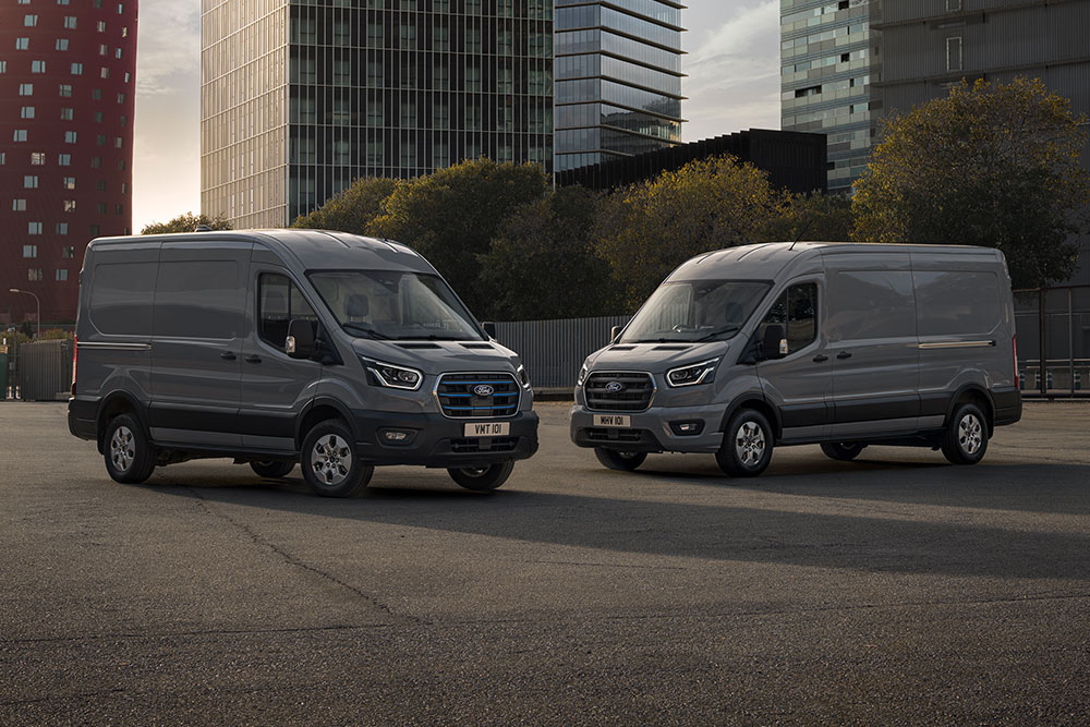 Two Ford Pro Transit vans, one of them electric, parked on a lot with modern buildings in the background.