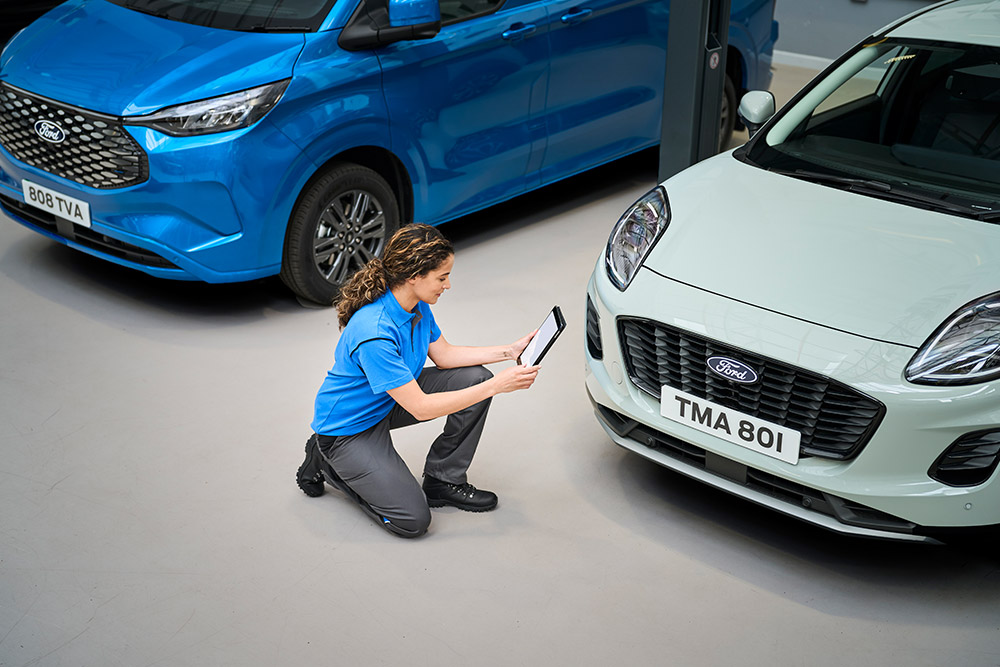 A technician kneeling between a blue Ford Transit Custom and a white Ford Puma, looking at a tablet.