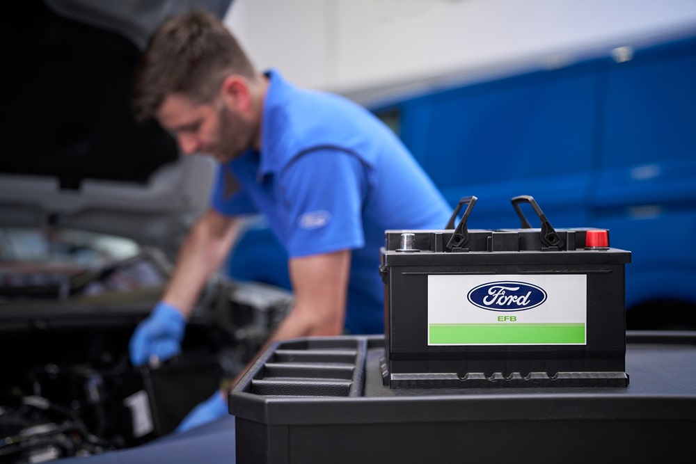 A Ford mechanic working on a car engine, with a Ford car battery in the foreground.