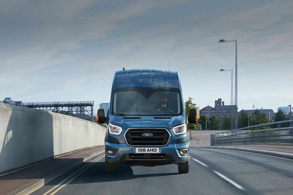 Front view of a blue Ford Transit van driving on a city road with a person at the wheel.