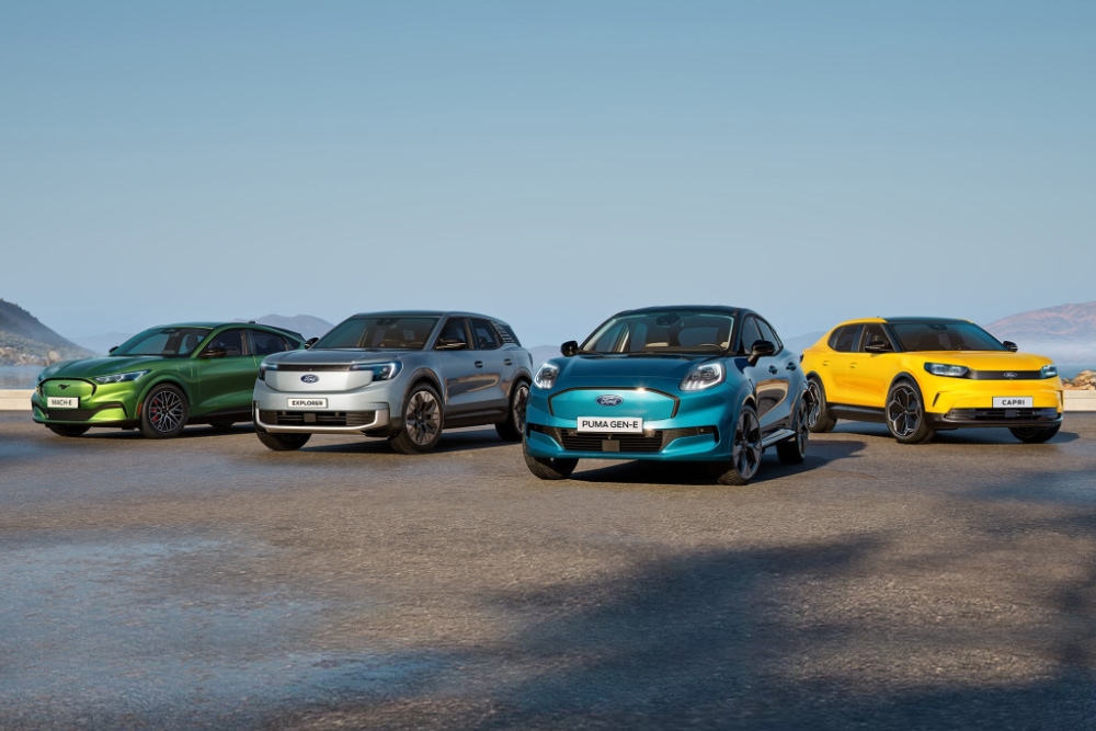 Four Ford electric vehicles: green Mach-E, silver Explorer, teal Puma Gen-E, and yellow Capri, on a coastal road.