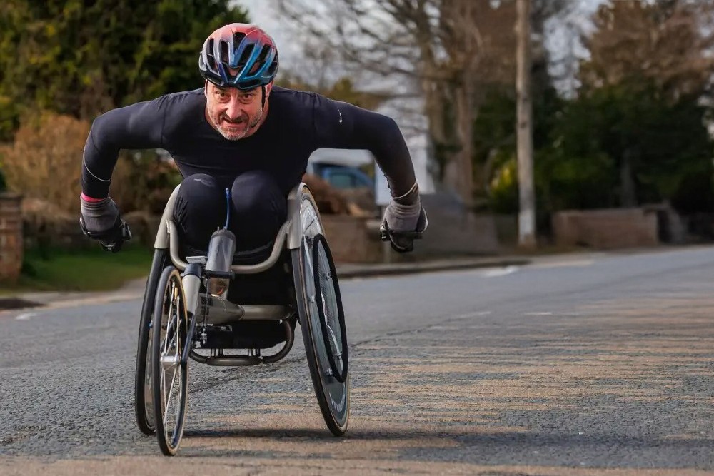 Jason Richards in a racing wheelchair wearing a helmet, propelling themselves along a road